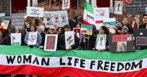 A group of protesters hold signs and banners during an Iranian solidarity demonstration. The front banner reads “Woman, Life, Freedom” in black letters on a green, white, and red background. Other signs reference Mahsa Amini, NIAC, and calls to boycott the Islamic Republic.