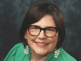 Headshot of an Indigenous woman. She has short dark brown hair, tortoise-shell glasses, is wearing red lipstick, a green shirt and white dangly earrings