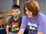 A middle-aged white woman with brown hair in a purple t-shirt cries. Next to her is a little boy with brown hair and a black t-shirt