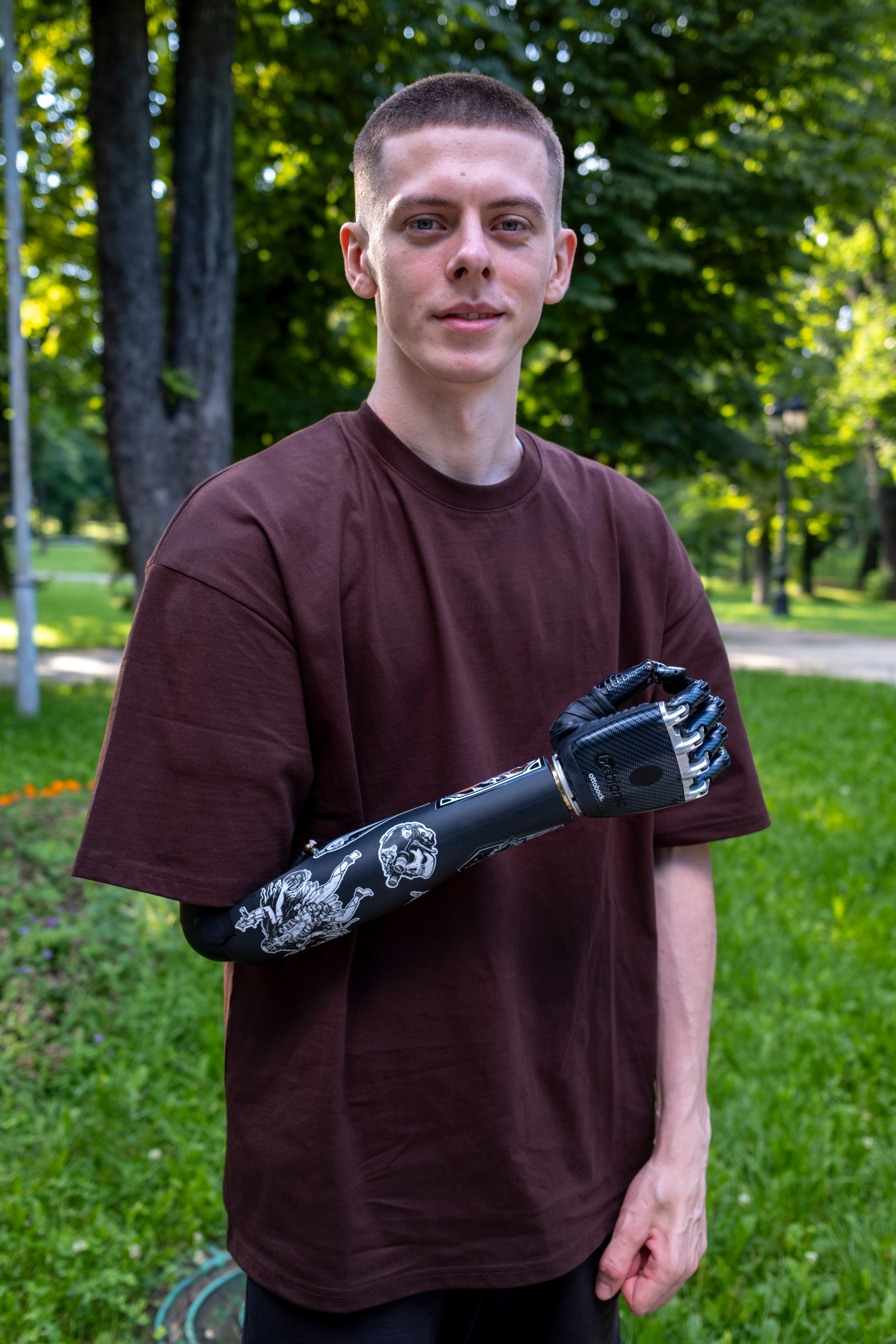 A young, white man with a buzz cut poses with his artificial arm. It is his right arm and made of black metal. He is wearing a brown t-shirt