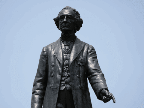 Image of a bronze statue of a man dressed in a petticoat with curls. He is pointing down with his index finger on his left hand