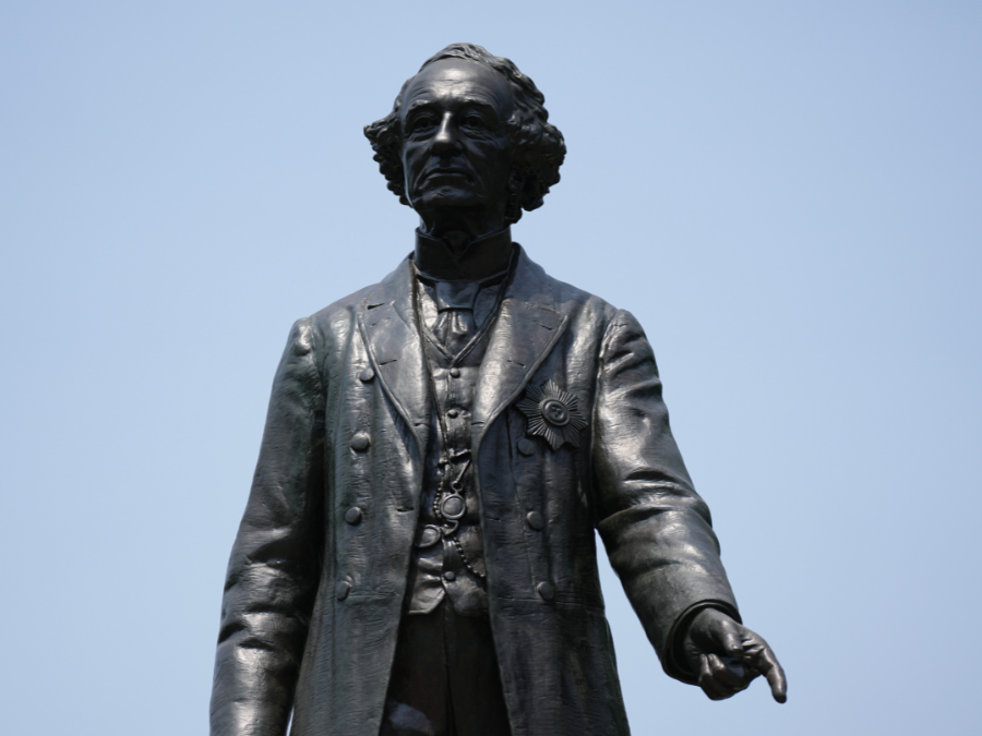 Image of a bronze statue of a man dressed in a petticoat with curls. He is pointing down with his index finger on his left hand
