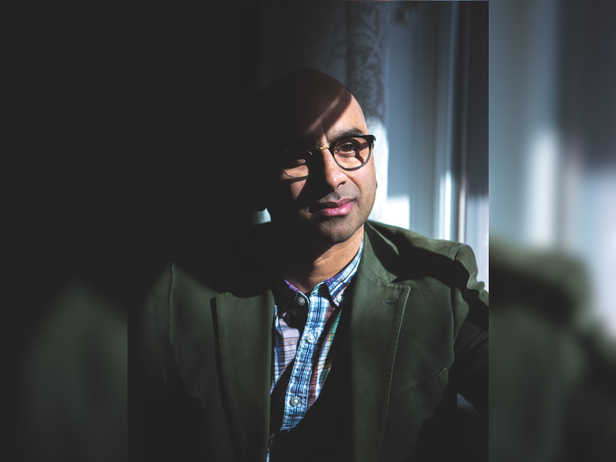 Headshot of a bald brown-skinned man with black-framed glasses. He is partially lit and there are shadows. He is wearing a blue checkered button-up shirt and dark coat
