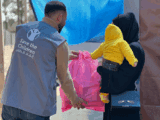 The back of an aid worker in a grey jacket as he hands a pink bag to a woman in black. She is holding a child in a yellow hoodie