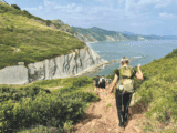 A group of people hike down a cliff. They are on a path and you can see greenery and water