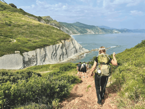 A group of people hike down a cliff. They are on a path and you can see greenery and water