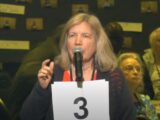 A Caucasian woman with blonde hair speaks at a microphone