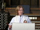 A Caucasian woman with shoulder-length red hair and minister's robes speaks at a pulpit