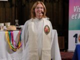A Caucasian woman with shoulder-length red hair and minister's robes stands at the front of a church