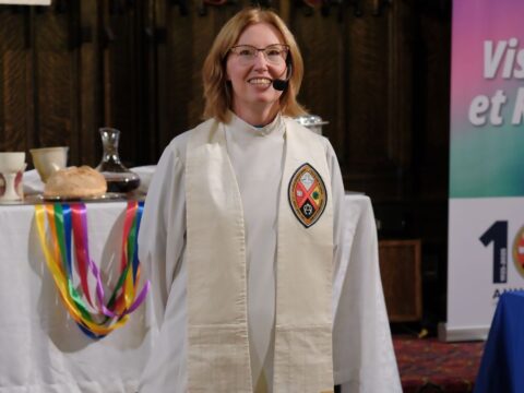 A Caucasian woman with shoulder-length red hair and minister's robes stands at the front of a church