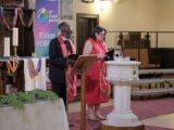 A Black man and an Indigenous woman wearing red stoles stand at the front of a church during a worship service.