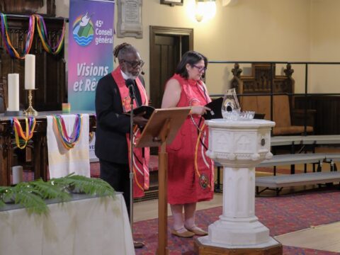 A Black man and an Indigenous woman wearing red stoles stand at the front of a church during a worship service.