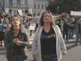 A young white woman with glasses and brown hair in a green coat and black shirt stands next to a white woman with brown hair. She is wearing a white cardigan and black shirt that says "FreePA". They are Christina nationalists in Elizabethtown, Pennsylvania