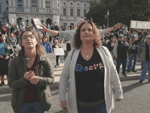A young white woman with glasses and brown hair in a green coat and black shirt stands next to a white woman with brown hair. She is wearing a white cardigan and black shirt that says "FreePA". They are Christina nationalists in Elizabethtown, Pennsylvania