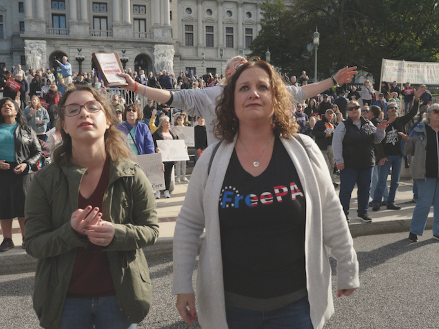 A young white woman with glasses and brown hair in a green coat and black shirt stands next to a white woman with brown hair. She is wearing a white cardigan and black shirt that says "FreePA". They are Christina nationalists in Elizabethtown, Pennsylvania