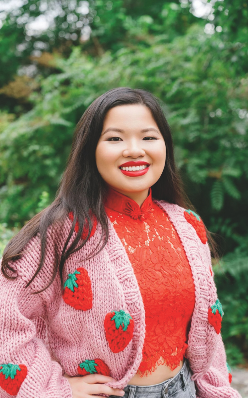 A young Asian woman with long black hair, red lipstick, a red shirt and pink cardigan with strawberries poses. She is smiling