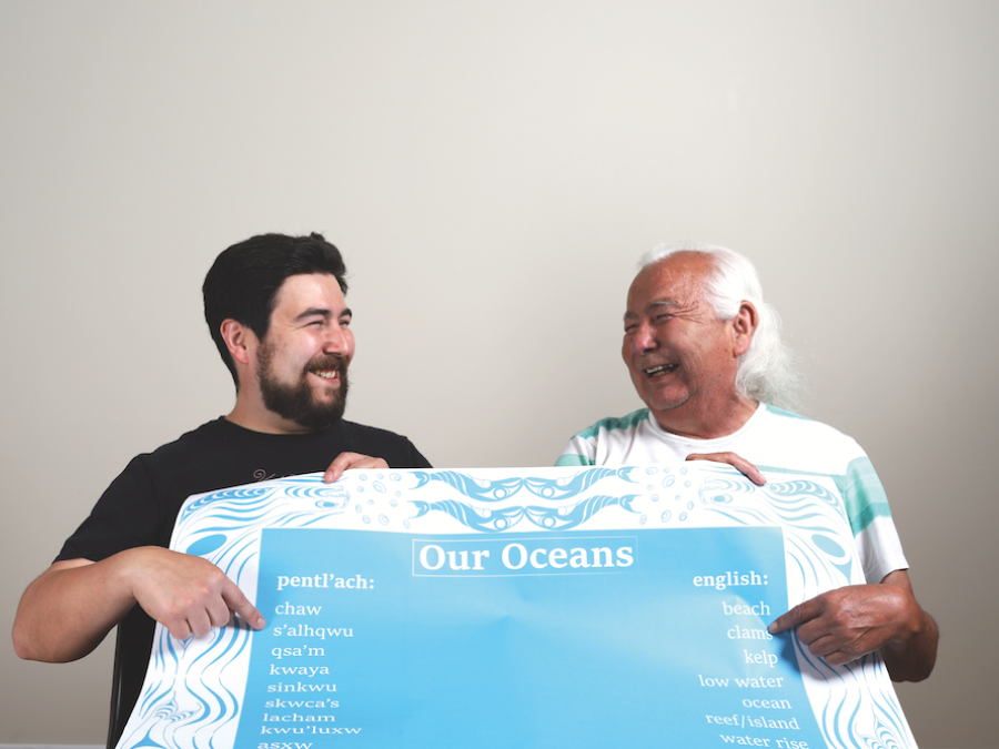 A young Indigenous man with dark brown hair and facial hair smiles and points to a sign he is holding up. Next to him is an Indigenous elder with white hair who is holding the other side of the sign and pointing at words. The sign says "Our Oceans" and has words in pentl'ach and English