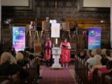 A Black man and an Indigenous woman wearing red stoles stand at the front of a church during a worship service.