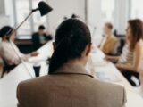 A woman wearing a brown coat is sitting on a chair in a boardroom with other colleagues.