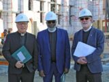 Three men, one east Asian, one Black and one Caucasian, stand wearing white hard hats and suit jackets at a construction site.
