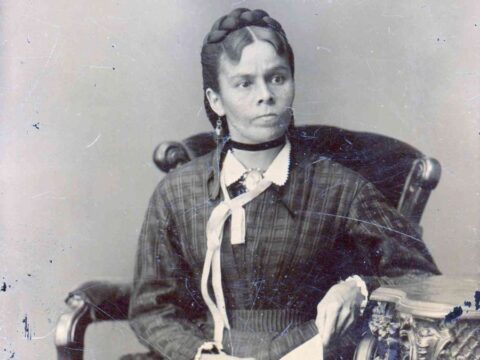 A black and white photo shows an Indigenous woman siting in a chair. Her hair is braided and pinned to her head and she is wearing a long dress.