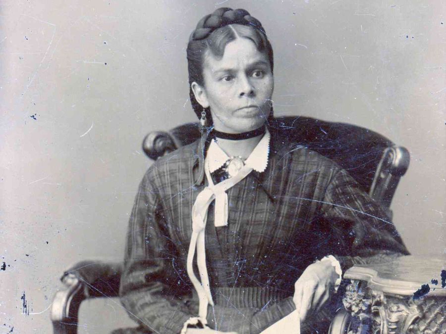 A black and white photo shows an Indigenous woman siting in a chair. Her hair is braided and pinned to her head and she is wearing a long dress.