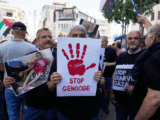 A man holding up a white poster with a red hand print that says stop genocide underneath.