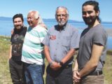 Four individuals, wearing casual clothes like jeans and a t-shirt, are standing outdoors near a scenic backdrop of a body of water and clear blue skies. The setting appears to be natural, with grass and a calm shoreline in view.