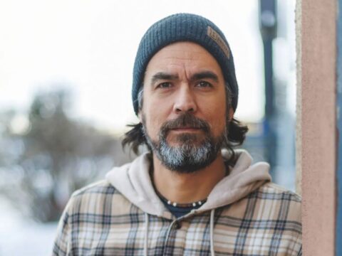 A person wearing a blue knit beanie, beige plaid button-up shirt over a coloured hoodie, standing outside near a building wall.