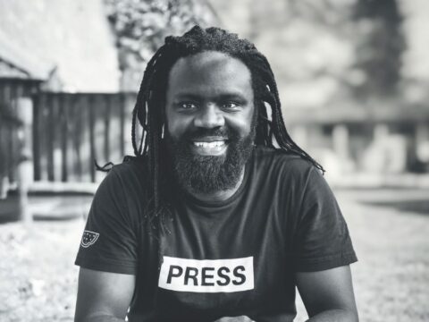 A man with long braided hair is sitting outdoors, wearing a black T-shirt with the word "PRESS" printed in bold letters on the front. The background features a blurred scene with a fence, trees, and open space.