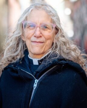 Elderly person with long gray hair and glasses smiles warmly, wearing a clerical collar and black jacket, standing outdoors with a blurred, colourful background.
