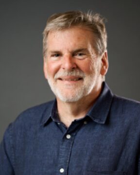 A smiling man with short gray hair and a beard, wearing a dark blue button-up shirt, poses against a plain gray background, conveying a friendly tone.