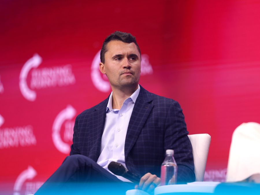A Caucasian man with brown hair in a blue suit jacket sits as he listens during a conference