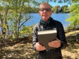 A man standing by the lake is smiling and holding a book.