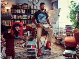 A person is seated confidently on a desk, surrounded by stacks of books on the floor, showcasing a scholarly or artistic environment