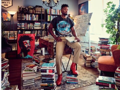 A person is seated confidently on a desk, surrounded by stacks of books on the floor, showcasing a scholarly or artistic environment