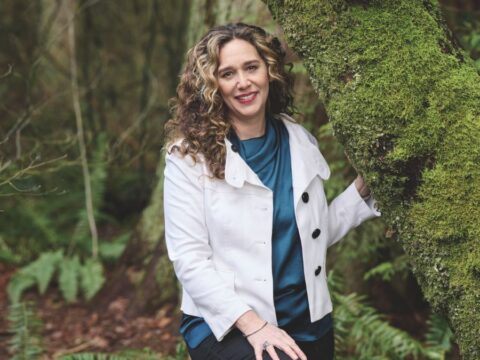 A woman in a white jacket and satin green shirt is standing and smiling beside a tree covered in moss.