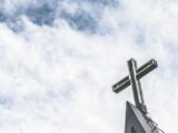 An image of a blueish grey sky with white wispy clouds and a church cross on the bottom right corner.