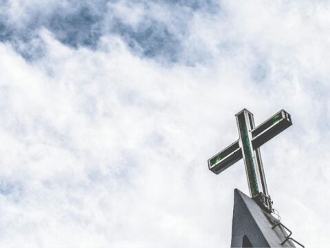 An image of a blueish grey sky with white wispy clouds and a church cross on the bottom right corner.