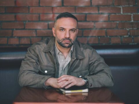 Man with short hair and a beard sits at a table in a dimly lit room, wearing a jacket. He looks thoughtful, with hands clasped over a small notebook.