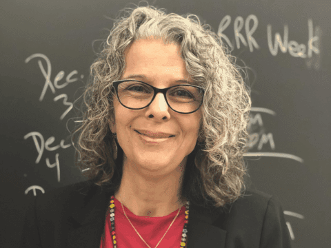 Smiling woman with curly gray hair and glasses stands in front of a chalkboard with writings. She wears a red top and colorful beaded necklace.
