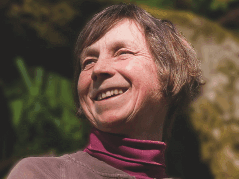 Smiling older woman with short hair, wearing a maroon turtleneck, stands outdoors in sunlight. Background shows blurred greenery and stone texture.