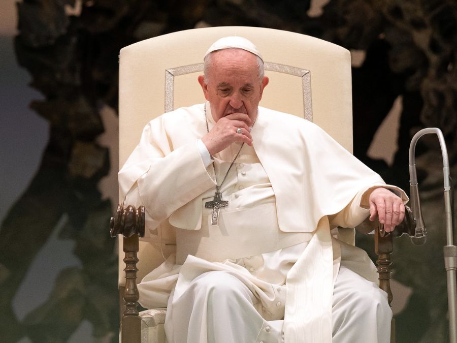 An elderly man in white robes and a hat sits in a chair