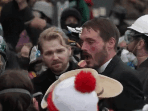 A crowded scene with people wearing protective gear and winter clothing. Two men stand out, one with a concerned expression, the other speaking passionately.