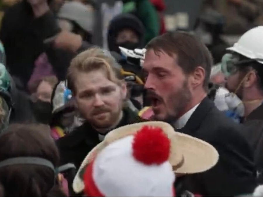 A crowded scene with people wearing protective gear and winter clothing. Two men stand out, one with a concerned expression, the other speaking passionately.