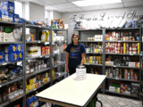 A woman smiles in a well-stocked food pantry with shelves filled with canned goods and boxes. A donation jar is on the table. Atmosphere is welcoming.
