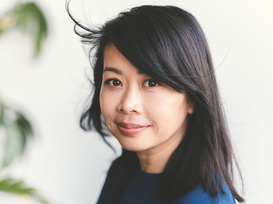 A woman with long, dark hair smiles softly towards the camera. She wears a navy blue top, with wisps of hair gently framing her face. The background is blurred with greenery.