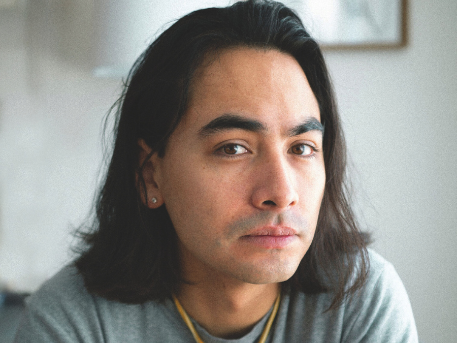 A person with long dark hair and a contemplative expression is seated indoors, dressed in a gray top. The lighting is soft, creating a calm and introspective mood.
