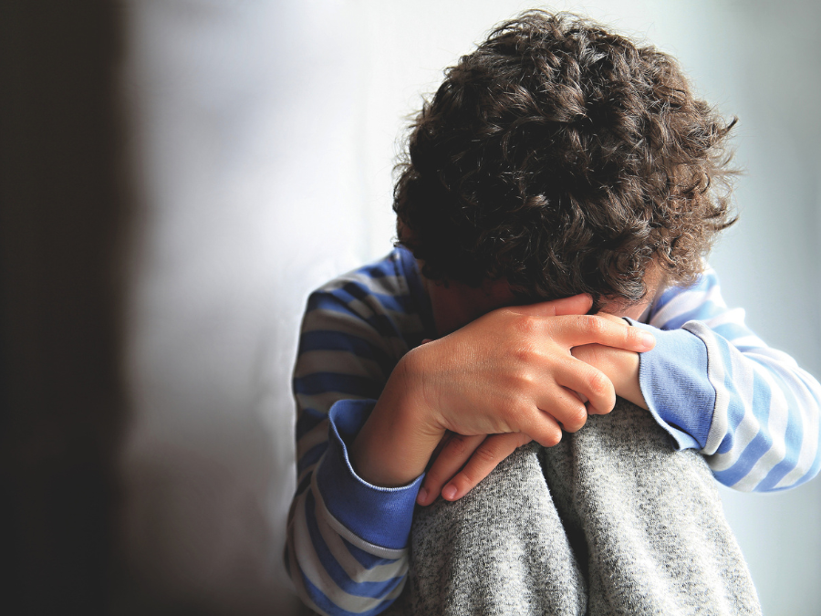 A young child with curly hair sits with head in hands, wearing a blue striped shirt and gray pants, conveying sadness and introspection in soft lighting.