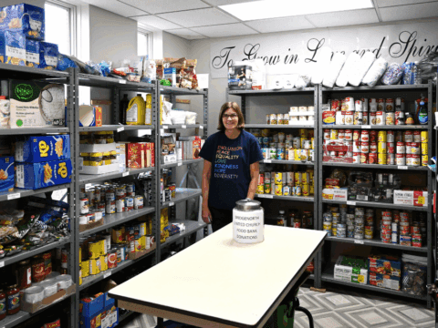 A woman smiles in a well-stocked food pantry with shelves filled with canned goods and boxes. A donation jar is on the table. Atmosphere is welcoming.